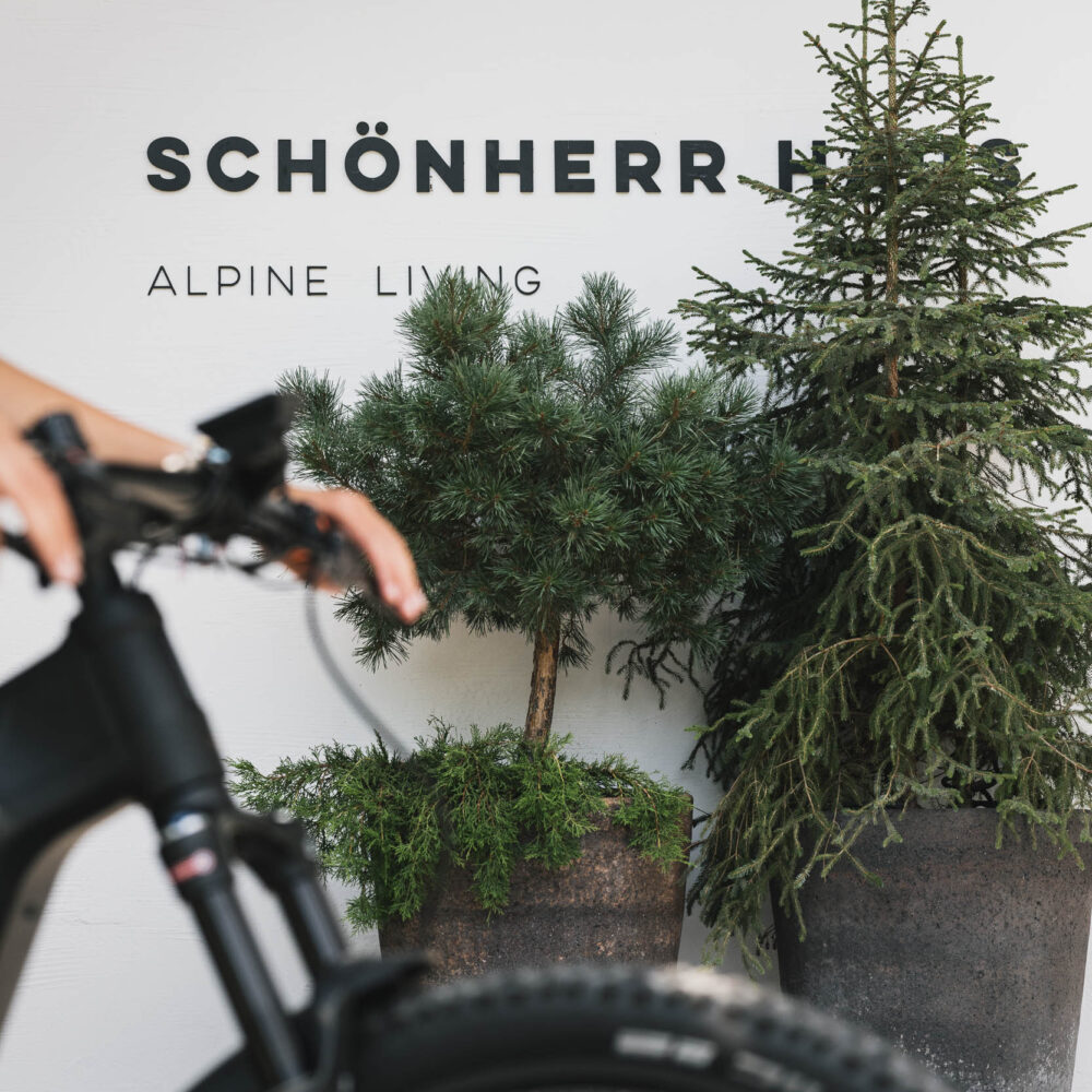 Schwarzes Mountainbike vor weißer Wand mit Schriftzug "SCHÖNHERR HAUS ALPINE LIVING" und mehreren Topfpflanzen.
